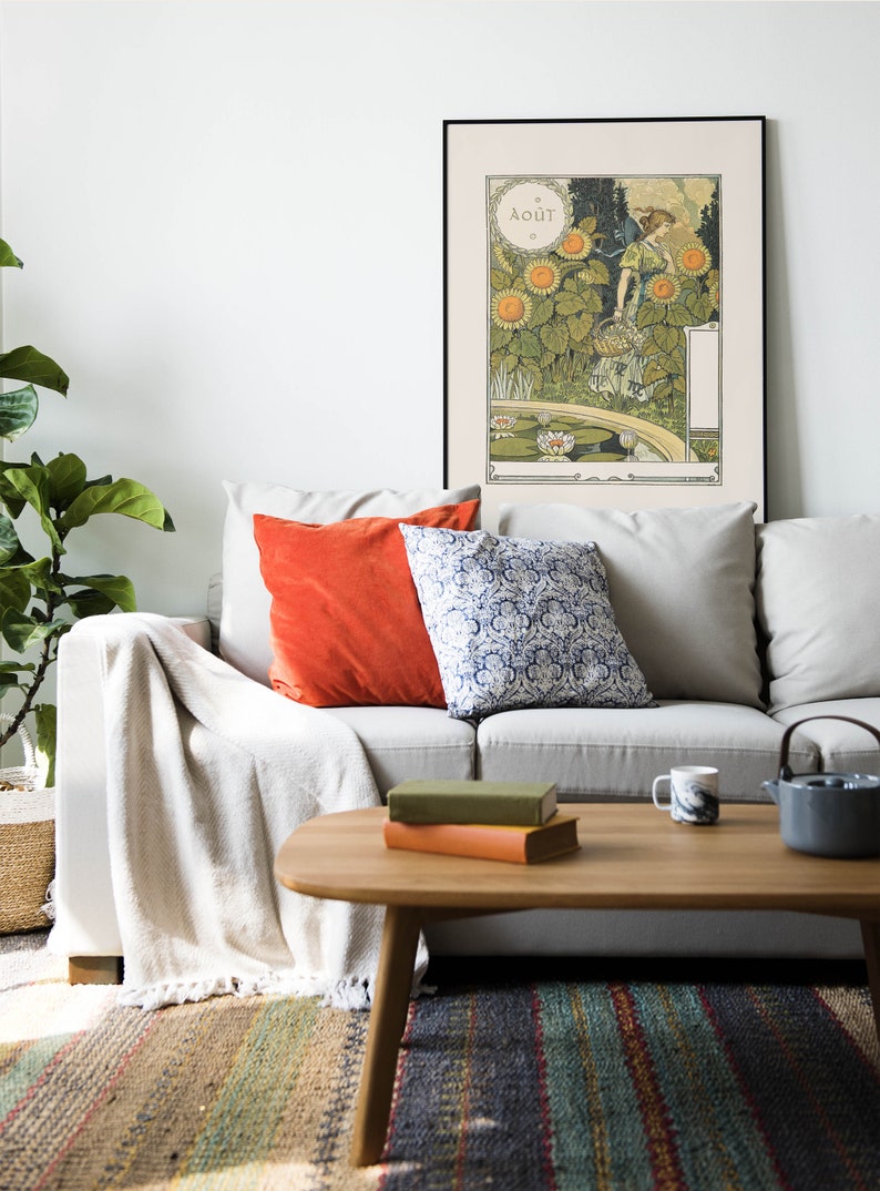 Puede incluir: Un sof&aacute; blanco con una manta, dos almohadas, una naranja y otra azul y blanca con estampado, se encuentra frente a una pared blanca con un p&oacute;ster enmarcado de una mujer en un campo de girasoles. El p&oacute;ster est&aacute; en franc&eacute;s y dice "Aout", que es la palabra francesa para agosto. El sof&aacute; est&aacute; frente a una mesa de centro con dos libros y una taza encima.