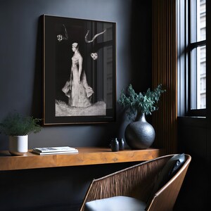May include: A black and white framed print of a woman in a long dress standing in a field. The print is hanging on a dark grey wall in a room with a wooden desk and a chair.