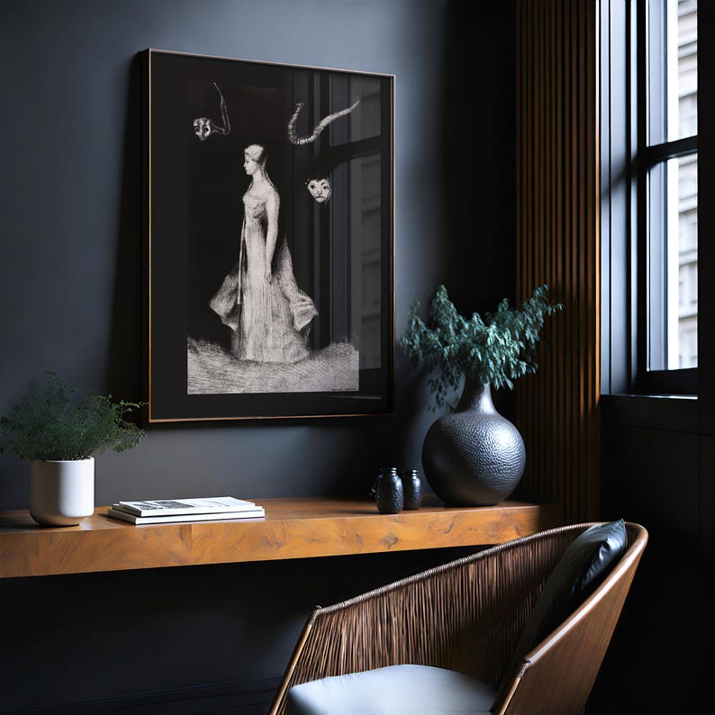May include: A black and white framed print of a woman in a long dress standing in a field. The print is hanging on a dark grey wall in a room with a wooden desk and a chair.