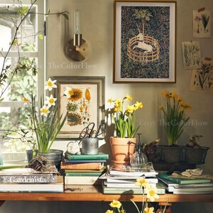 以下が含まれることがあります： 本、鉢植えの水仙、額入りのアートワークが飾られた木製のテーブルがある居心地の良いインテリアシーン。アートワークには植物画や風景画が含まれています。壁の燭台と窓が部屋の雰囲気を高めています。「Gardenista」というテキストが見えます。