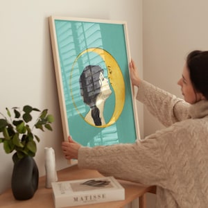 Puede incluir: Una impresi&oacute;n enmarcada con un fondo azul verdoso que presenta un rostro de mujer de perfil besando una luna creciente. La luna es amarilla y el cabello de la mujer es negro. La impresi&oacute;n est&aacute; en un marco de madera.