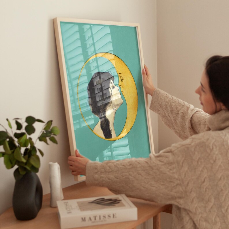 Puede incluir: Una impresi&oacute;n enmarcada con un fondo azul verdoso que presenta un rostro de mujer de perfil besando una luna creciente. La luna es amarilla y el cabello de la mujer es negro. La impresi&oacute;n est&aacute; en un marco de madera.