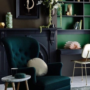 Puede incluir: Un sill&oacute;n de terciopelo verde oscuro con un coj&iacute;n beige claro se encuentra en una habitaci&oacute;n con paredes verde oscuro y una chimenea negra. Una mesa peque&ntilde;a con una tapa de m&aacute;rmol blanco se encuentra frente al sill&oacute;n.