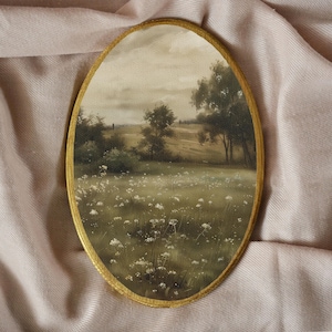 May include: Oval landscape painting in a gold frame. The artwork shows a field of white wildflowers, trees, and a cloudy sky. The painting has a vintage aesthetic. The background is a soft, pink fabric.