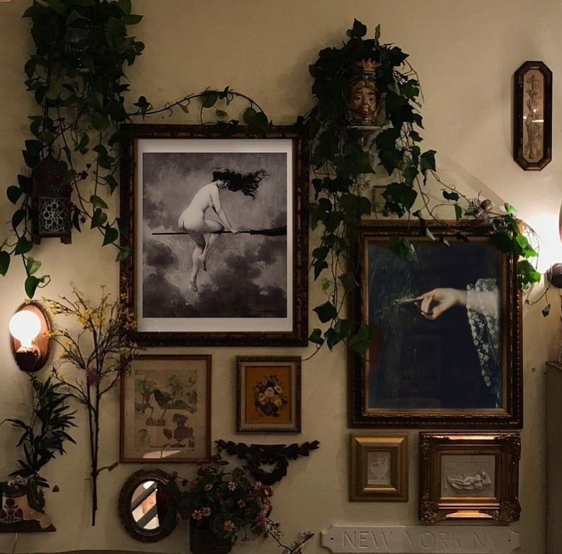 May include: A wall gallery featuring a variety of framed artwork, including a black and white illustration of a woman riding a broomstick, a painting of a hand holding a feather, and a framed print of a floral design. The gallery is decorated with greenery and a small light fixture.