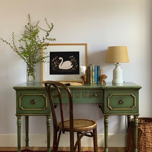 May include: A green painted wooden desk with a gold trim and two drawers. A woven chair sits in front of the desk. A framed print of a white swan on a black background hangs on the wall above the desk. A lamp with a beige shade sits on the desk.