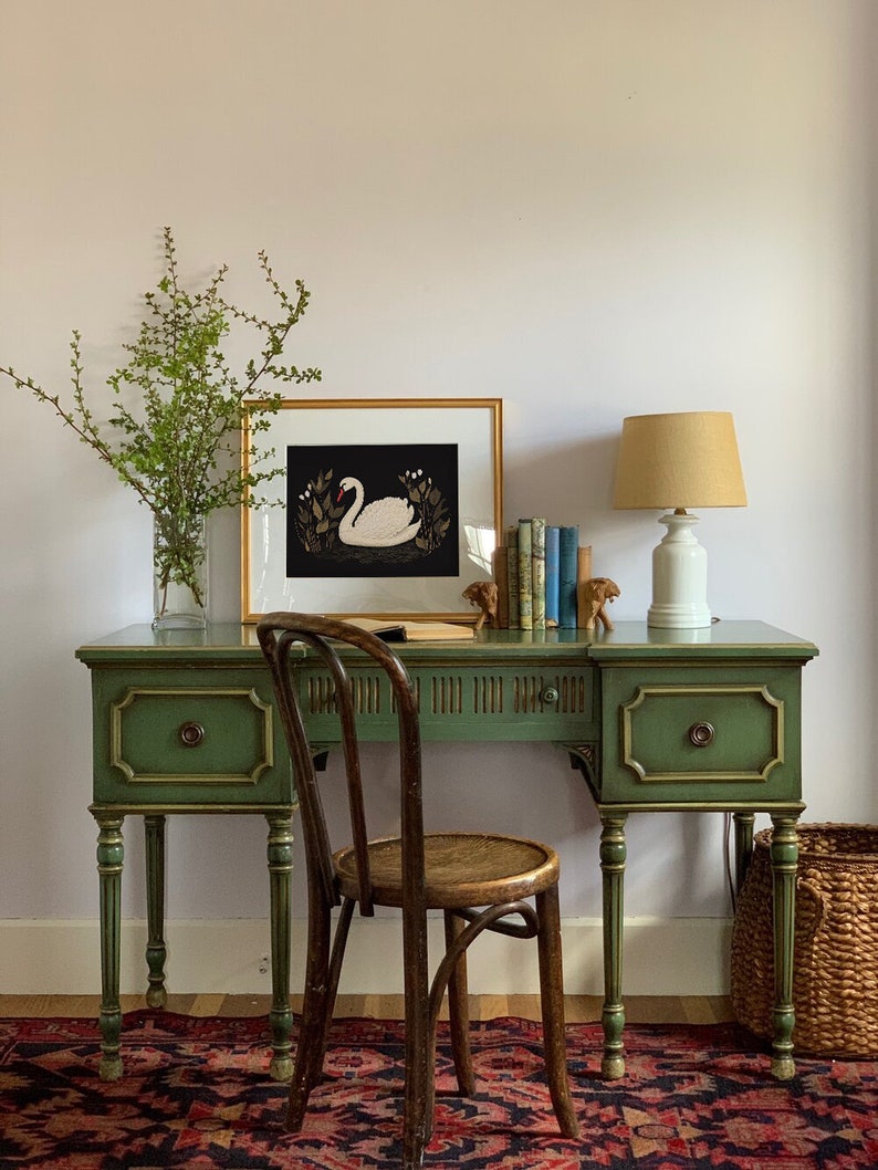 May include: A green painted wooden desk with a gold trim and two drawers. A woven chair sits in front of the desk. A framed print of a white swan on a black background hangs on the wall above the desk. A lamp with a beige shade sits on the desk.