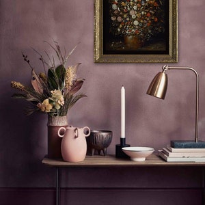 Peut inclure: Une peinture de nature morte repr&eacute;sentant un vase rempli de fleurs color&eacute;es, encadr&eacute;e d'or et accroch&eacute;e &agrave; un mur mauve. Une table avec un vase rose, une bougie, un bol et des livres se trouve devant la peinture.