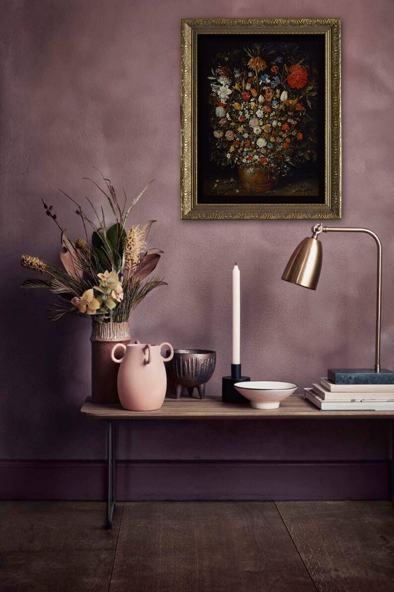Peut inclure: Une peinture de nature morte repr&eacute;sentant un vase rempli de fleurs color&eacute;es, encadr&eacute;e d'or et accroch&eacute;e &agrave; un mur mauve. Une table avec un vase rose, une bougie, un bol et des livres se trouve devant la peinture.