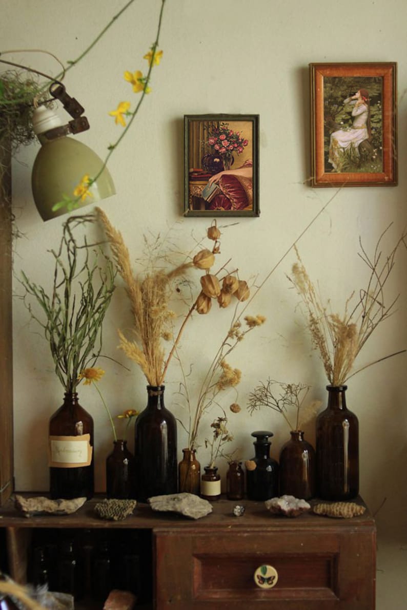May include: A collection of dried flowers and grasses in brown glass bottles arranged on a wooden shelf with a drawer. The shelf has a small butterfly design on the drawer. Two framed pictures hang on the wall behind the shelf.