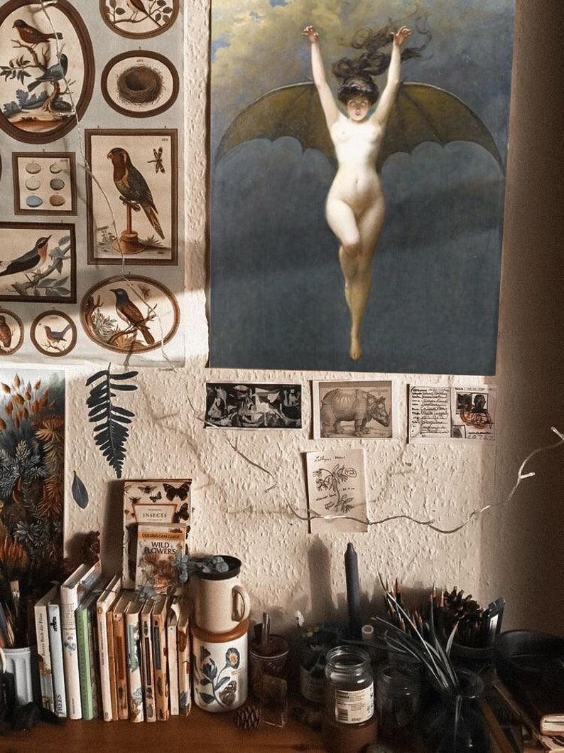 Op de afbeelding: Een witte muur met een ingelijst schilderij van een gevleugelde vrouw, een varen en een plank met boeken, een mok en kaarsen. Het schilderij is in vintage stijl en de vrouw is naakt. De plank is versierd met verschillende items, waaronder boeken, een mok en kaarsen.