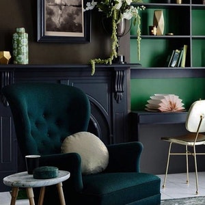 May include: A dark green velvet armchair with a light beige pillow sits in a room with a dark green and black fireplace and a dark green bookshelf. The room is decorated with a black and white framed print of a woman on a broomstick.