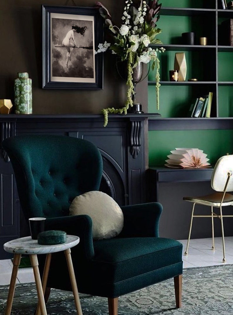 May include: A dark green velvet armchair with a light beige pillow sits in a room with a dark green and black fireplace and a dark green bookshelf. The room is decorated with a black and white framed print of a woman on a broomstick.