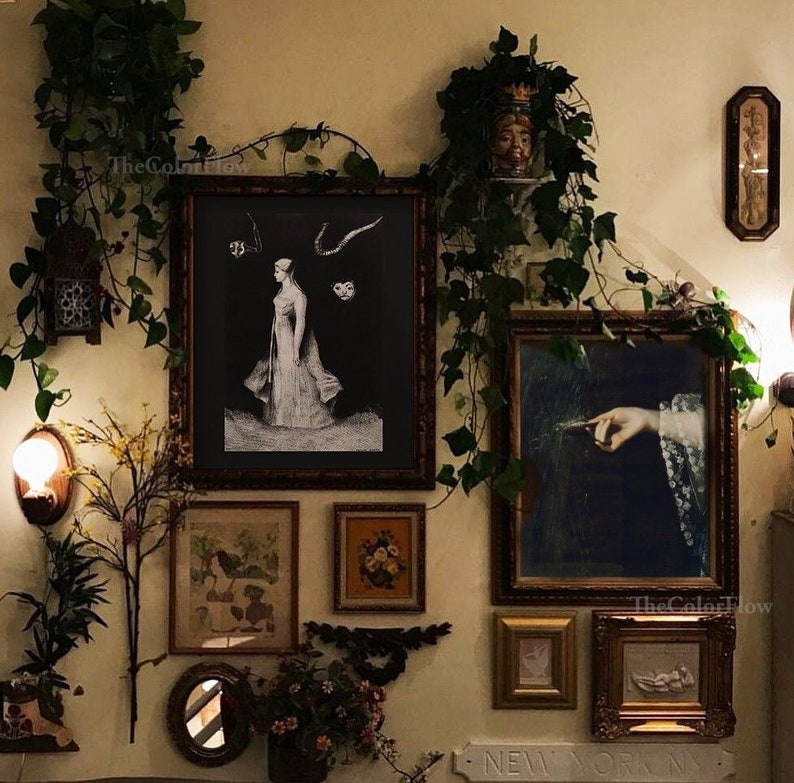 May include: A wall decorated with framed artwork, including a black and white illustration of a woman in a flowing dress, a painting of a hand holding a feather, and a floral print. The wall is adorned with greenery and a small potted plant.