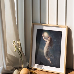 May include: A framed print of a woman holding a glowing orb in a dark sky. The print is on a wooden floor with a gold tray, a small white vase with white flowers, and a wooden sphere.