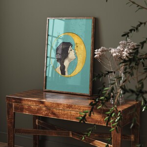 Puede incluir: Una impresi&oacute;n enmarcada de una mujer con cabello largo y oscuro besando una luna creciente. La luna es amarilla y el fondo es turquesa.