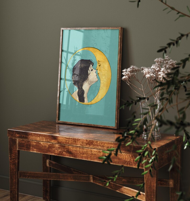 Puede incluir: Una impresi&oacute;n enmarcada de una mujer con cabello largo y oscuro besando una luna creciente. La luna es amarilla y el fondo es turquesa.