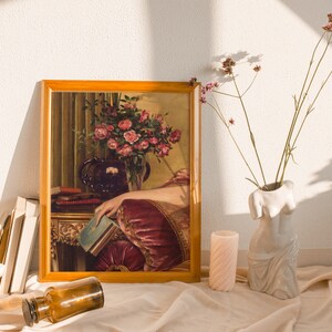 May include: A framed painting of a woman in a burgundy dress holding a book. The painting is set on a table with a vase of pink roses and a stack of books. The painting is surrounded by a wooden frame.