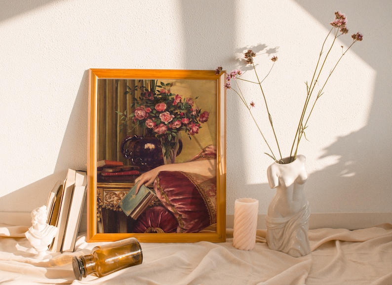 May include: A framed painting of a woman in a burgundy dress holding a book. The painting is set on a table with a vase of pink roses and a stack of books. The painting is surrounded by a wooden frame.