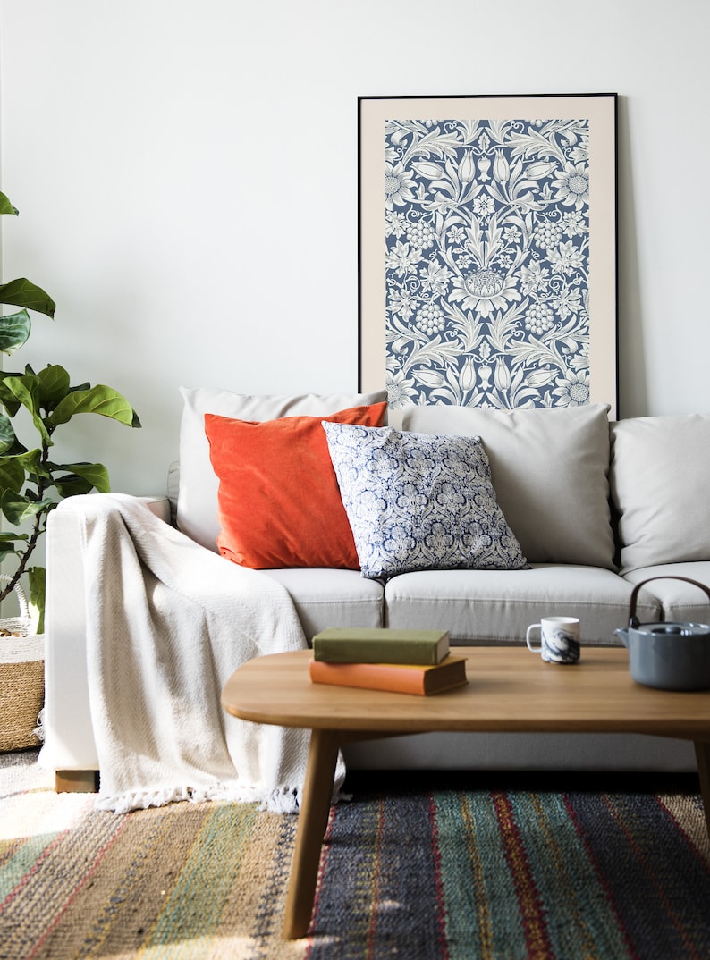 May include: A white couch with a throw blanket and two patterned pillows. A framed print with a blue and white floral pattern hangs on the wall above the couch. A wooden coffee table with books and a mug sits in front of the couch.