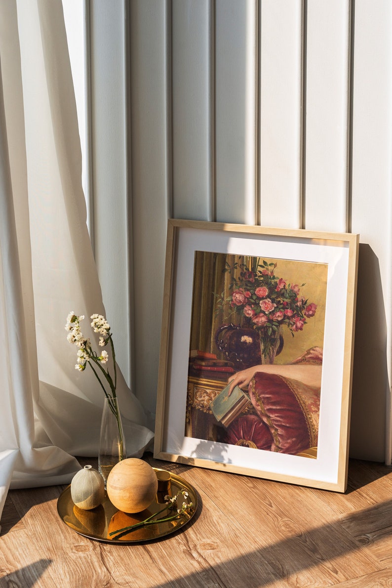 May include: A framed painting of a woman in a red dress holding a book, with a vase of pink roses in the background. The painting is on a wooden floor with a gold tray, two decorative spheres, and a vase of white flowers.