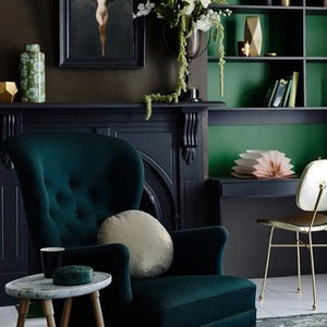 Op de afbeelding: Een donkergroene fluwelen fauteuil met een gouden kussen staat in een kamer met een donkergroen en bruin kleurenschema. Een zwarte open haard met een gouden decoratief element bevindt zich achter de fauteuil. Een kleine ronde tafel met een wit marmeren blad staat voor de fauteuil.