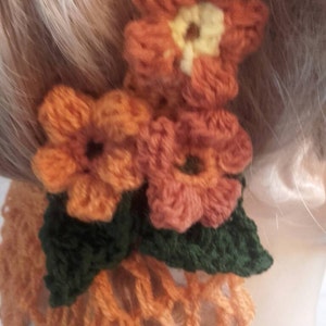 May include: A crocheted hair scrunchie with a brown and orange color scheme. The scrunchie features three crocheted flowers and green leaves.