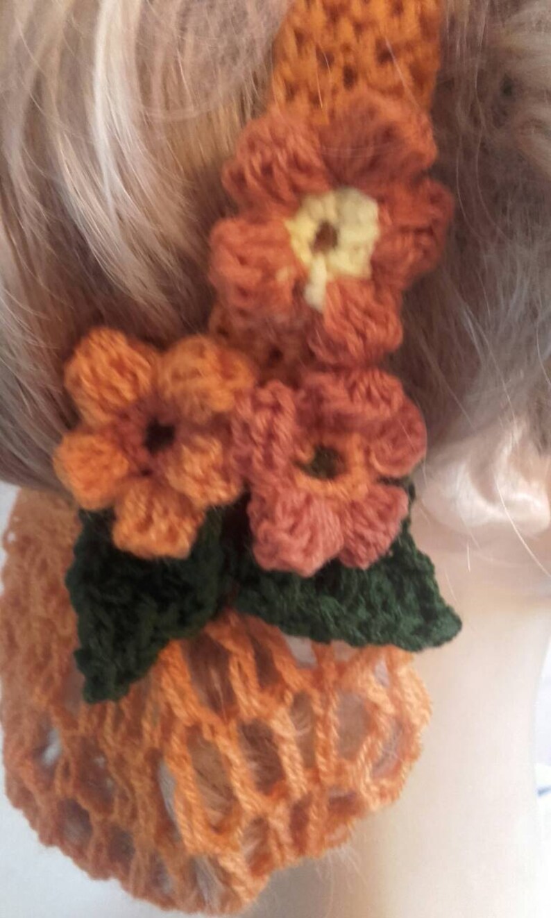 May include: A crocheted hair scrunchie with a brown and orange color scheme. The scrunchie features three crocheted flowers and green leaves.