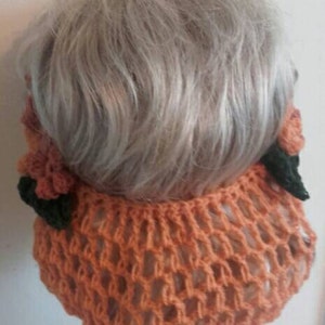May include: A crocheted hairnet with a peach color and floral accents. The hairnet is made of a delicate, open-weave material and features two small, crocheted flowers on each side.