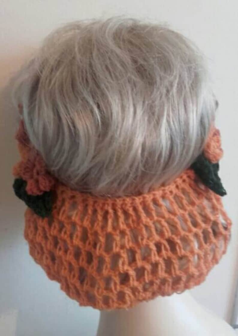 May include: A crocheted hairnet with a peach color and floral accents. The hairnet is made of a delicate, open-weave material and features two small, crocheted flowers on each side.