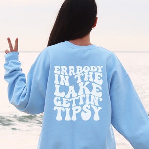 May include: A light blue sweatshirt with the text "ERRBODY IN THE LAKE GETTIN' TIPSY" in white, retro-style lettering. The sweatshirt is seen from the back, with a person standing near the water.