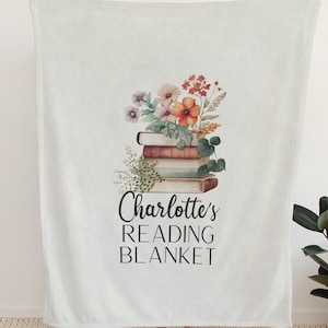 May include: A white fleece blanket with a floral design and a stack of books. The text "Charlotte's Reading Blanket" is printed on the blanket.