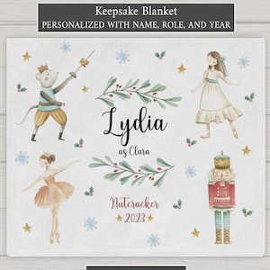 May include: A personalized keepsake blanket featuring a watercolor illustration of a Nutcracker, a ballerina, and a mouse. The blanket is customized with the name "Lydia" and the role "Clara" for the Nutcracker performance. The year "2023" is also included. The blanket is available in minky or sherpa fabric.