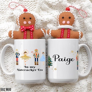 Puede incluir: Dos tazas blancas de 15 oz con un tema de Cascanueces. Una taza tiene el texto "In my Nutcracker Era" y la otra taza tiene el nombre "Paige". Ambas tazas tienen un adorno de hombre de jengibre en la parte superior.