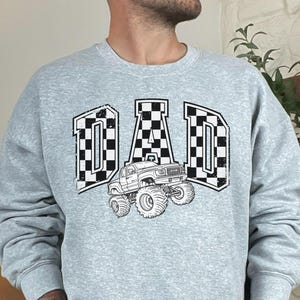 May include: Sport grey sweatshirt with the word "DAD" in a checkered flag design, with a monster truck graphic. The sweatshirt is a crew neck style.