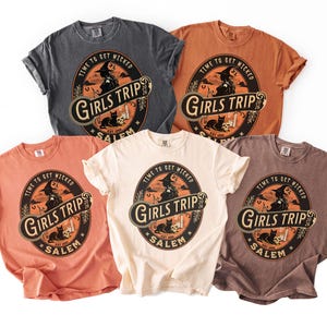 May include: Five short-sleeved crew neck t-shirts in charcoal, orange, peach, cream, and brown. Each shirt features a circular graphic with a witch, a black cat, and the text "TIME TO GET WICKED", "GIRLS TRIP", "SALEM", and "2026" in a vintage style.