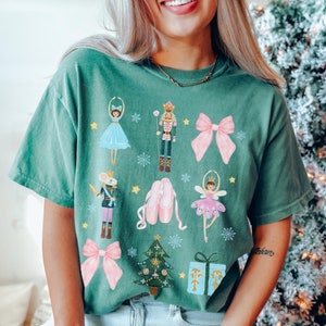 May include: A light green t-shirt with a festive Christmas design featuring a nutcracker, ballerina, mouse king, and other holiday icons. The design is in a watercolor style with pink, blue, and gold accents.