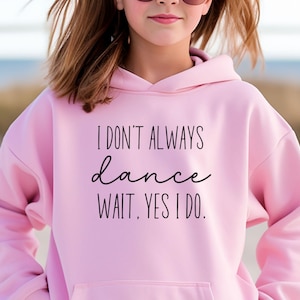 May include: A light pink hoodie with the text "I DON'T ALWAYS dance WAIT, YES I DO." in black script. The hoodie has a hood and a front pocket. The image shows a person wearing the hoodie.