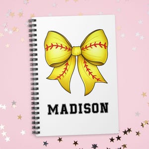 May include: A spiral-bound notebook with a white cover featuring a large yellow bow design with red stitching, resembling a softball. The name "MADISON" is printed in bold, black letters below the bow. The background is pink with star-shaped confetti.