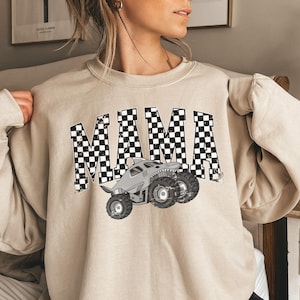 May include: Beige sweatshirt with the word "MAMA" in a checkered pattern. A gray monster truck with shark teeth is printed below the word. The sweatshirt has long sleeves and a crew neck.