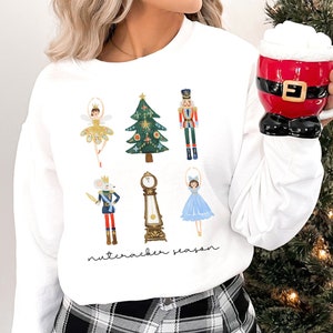 May include: White sweatshirt with a festive Nutcracker theme. The design features a Christmas tree, a Nutcracker soldier, a ballerina, a mouse king, a clock, and the text "Nutcracker Season".