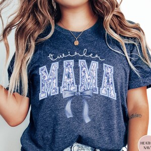 May include: Heather navy blue t-shirt with the words "twirlee MAMA" in white script and blue floral print. A blue bow is centered below the word "MAMA". The shirt is a crew neck style.