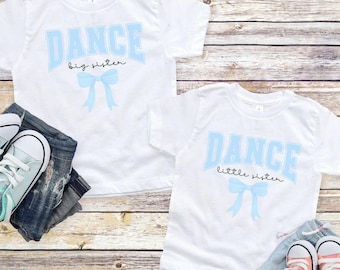 Puede incluir: Dos camisetas blancas con la palabra "DANCE" en letras de bloque azul claro. Una camiseta dice "big sister" y la otra "little sister" en cursiva. Ambas camisetas tienen un lazo azul claro. Vaqueros y zapatillas deportivas están junto a las camisetas.