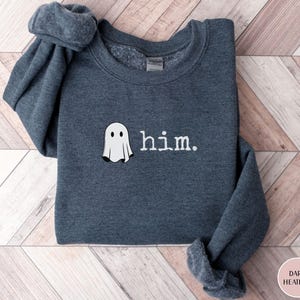 May include: A dark heather gray crewneck sweatshirt featuring an embroidered white ghost graphic positioned to the left of the lowercase white text that reads him. The garment has long sleeves and a ribbed collar, cuffs, and hemline.