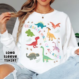 May include: White long-sleeve t-shirt featuring colorful dinosaur illustrations and the text "CELEBRATE MINDS OF ALL KINDS." The design includes various dinosaurs in green, red, blue, yellow, and purple, with fern accents.