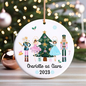 May include: A white round Christmas ornament with a watercolor illustration of a Nutcracker, two ballerinas, a mouse king, and a Christmas tree. The ornament is personalized with the text "Charlotte as Clara 2023".