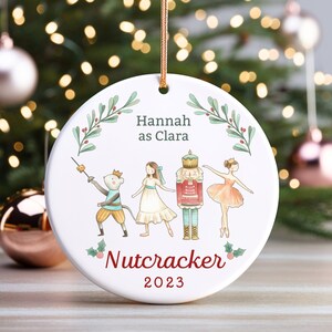 May include: White ceramic ornament with a watercolor illustration of characters from the Nutcracker ballet. The ornament features a mouse king, Clara, the Nutcracker, and a ballerina. The text "Hannah as Clara" and "Nutcracker 2023" is printed on the ornament.