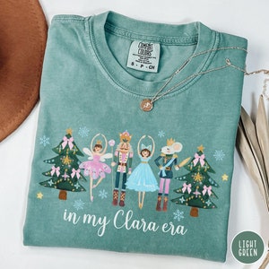 May include: A light green t-shirt with a festive design featuring a nutcracker, a ballerina, a mouse king, and two Christmas trees. The text "in my Clara era" is printed below the design.
