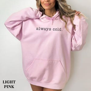 May include: Light pink hoodie with the text "always cold." The hoodie has a drawstring hood and a front pocket. The words "LIGHT PINK" are at the bottom of the image.