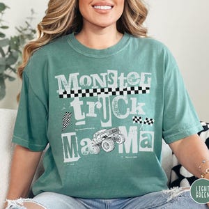 May include: A light green t-shirt with the words "Monster Truck Mama" in a distressed white font. The design includes a monster truck graphic and checkered flag accents. The shirt is a relaxed fit.